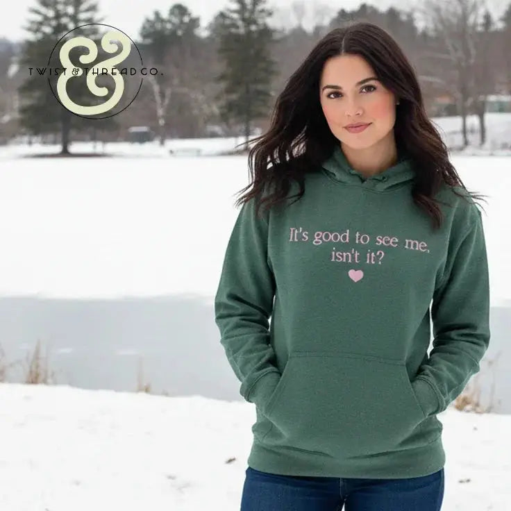 Woman wearing green hoodie with pink text 'It's good to see me, isn't it?' outdoors in snowy winter setting