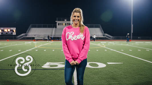Woman in pink 'Cheer' sweatshirt standing on football field at night, Twist & Thread Co. logo