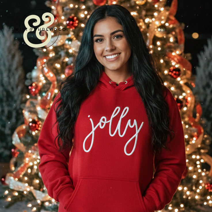 Smiling woman wearing red hoodie with 'jolly' text in front of decorated Christmas tree with lights and ornaments