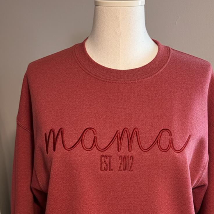 Pink folded sweatshirt with red embroidery saying 'mama EST. 2020' and side sleeve text with a heart