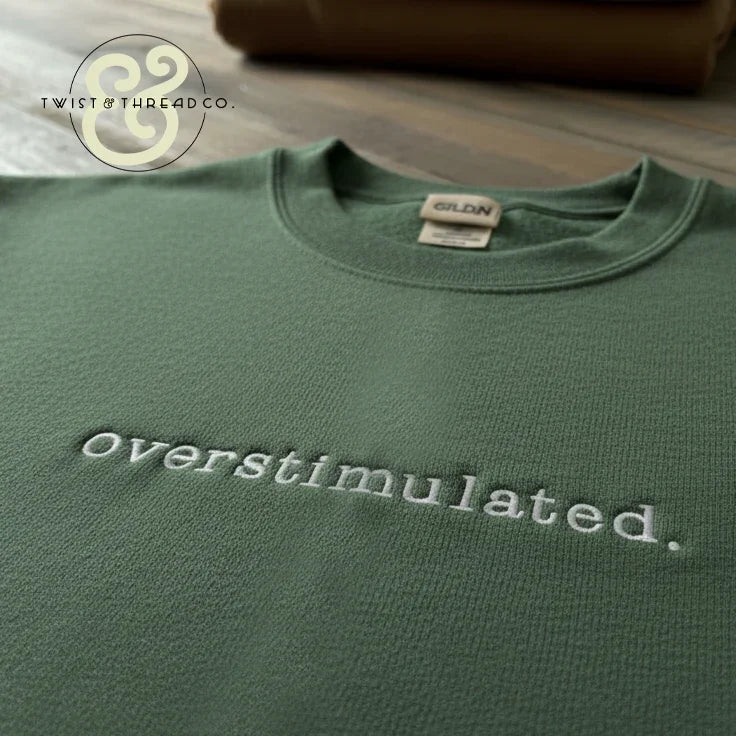 Green Gildan sweatshirt with embroidered text 'Overstimulated.' on wooden surface, Twist & Thread Co. logo