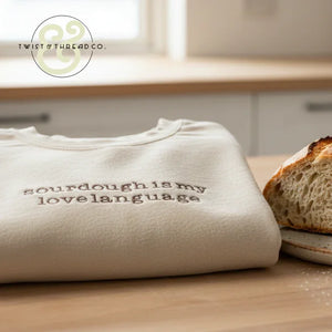 Folded cream-colored shirt with embroidered text 'sourdough is my lovelanguage' beside a slice of crusty sourdough bread on wooden table