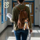 Teacher walking in school hallway wearing brown backpack with colorful teacher-themed bag tag and holding papers