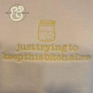Embroidered beige fabric with yellow text 'just trying to keep this bitch alive' and jar design from Twist & Thread Co.