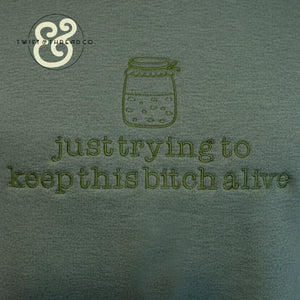 Green fabric with embroidered jar and text 'just trying to keep this bitch alive' by Twist & Thread Co.