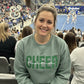 Smiling woman wearing green CHEER mama sweatshirt at a busy cheerleading competition arena