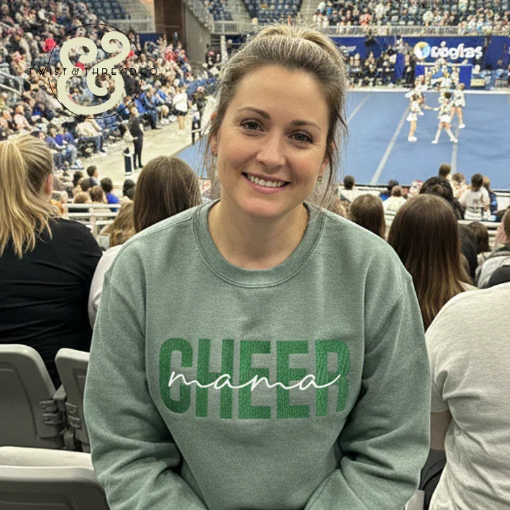 Smiling woman wearing green CHEER mama sweatshirt at a busy cheerleading competition arena