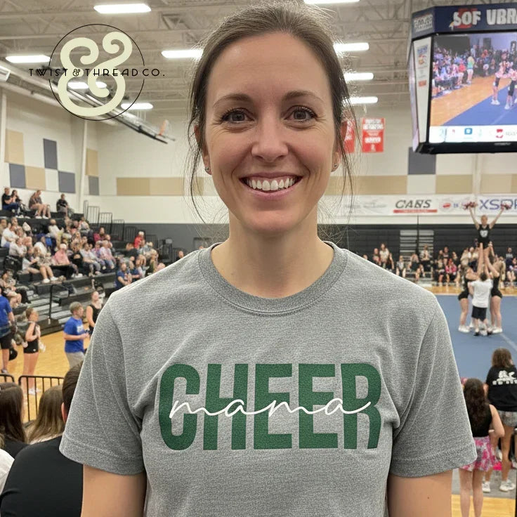 Woman smiling in gray Cheer Mama t-shirt at indoor cheerleading event with crowd and cheerleaders