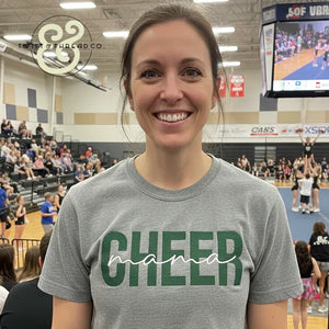 Woman smiling in gray Cheer Mama t-shirt at indoor cheerleading event with crowd and cheerleaders