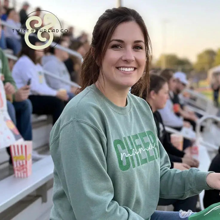 Smiling woman wearing green Cheer Mama embroidered sweatshirt sitting in stadium bleachers at outdoor event