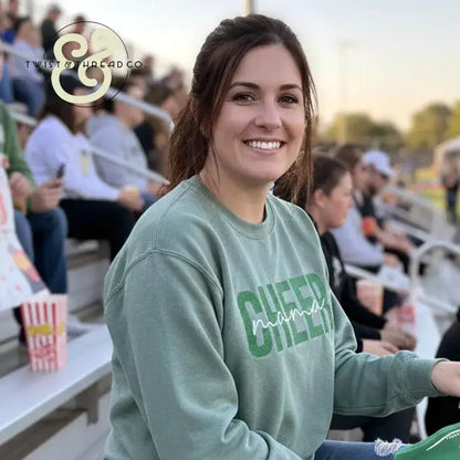 Smiling woman wearing green Cheer Mama embroidered sweatshirt sitting in stadium bleachers at outdoor event