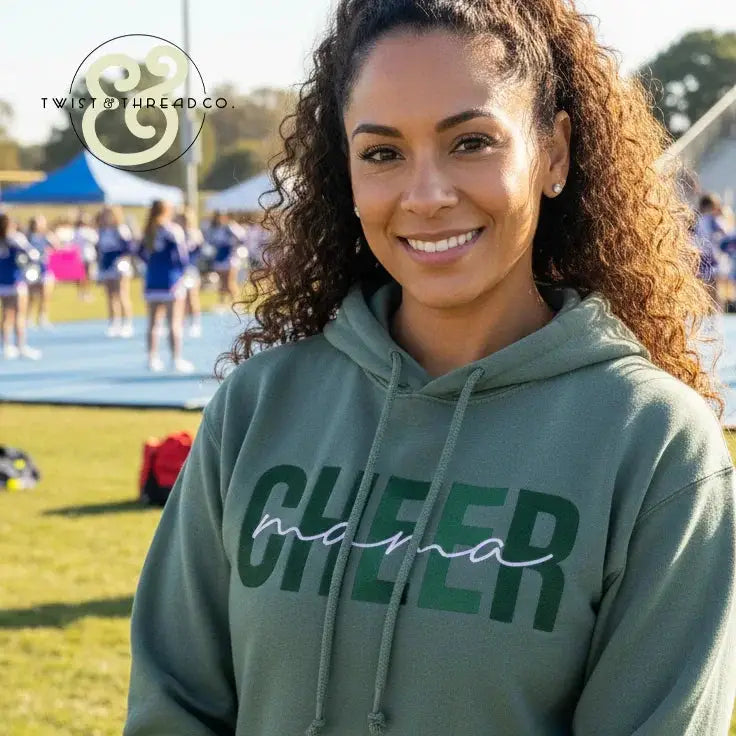 Smiling woman wearing green hoodie with embroidered CHEER mama text at outdoor cheerleading event