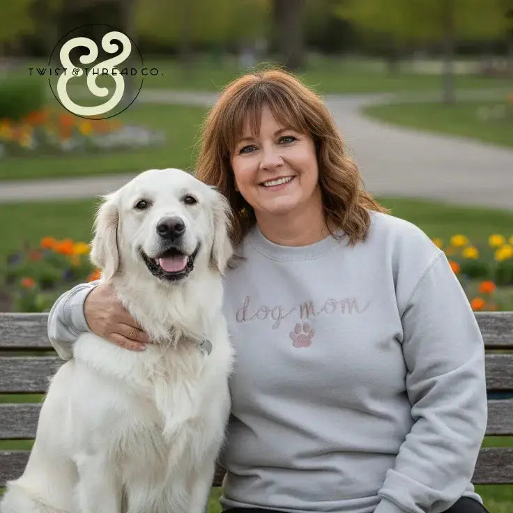 Smiling woman in 'dog mom' embroidered sweatshirt with white dog on park bench, Twist & Thread Co.