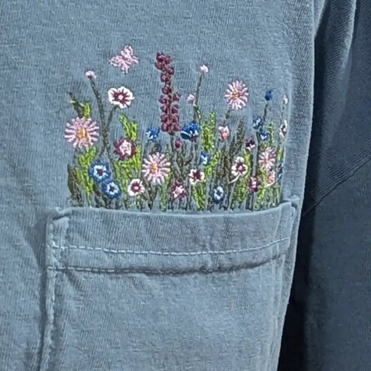 A delicate embroidered floral patch in soft pastel hues of pink, blue, and purple adorns the front pocket of a denim jacket.