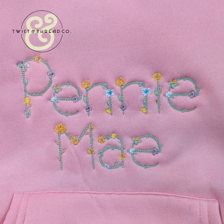 Pink fabric with embroidered floral text reading Penny Mae, premium custom thread work