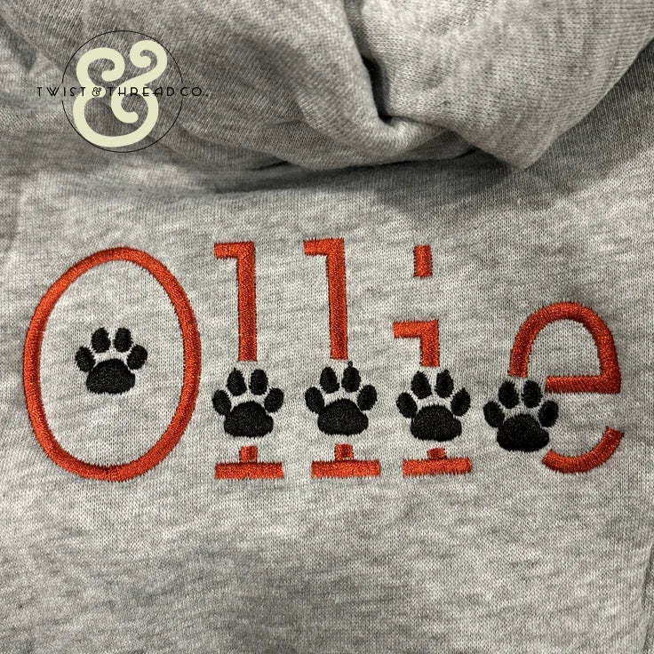 Gray hoodie with custom embroidered name 'Ollie' in orange and black paw prints at Twist & Thread Co.