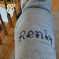 Gray dog hoodie with embroidered name 'Renly' and black paw prints, wooden staircase background