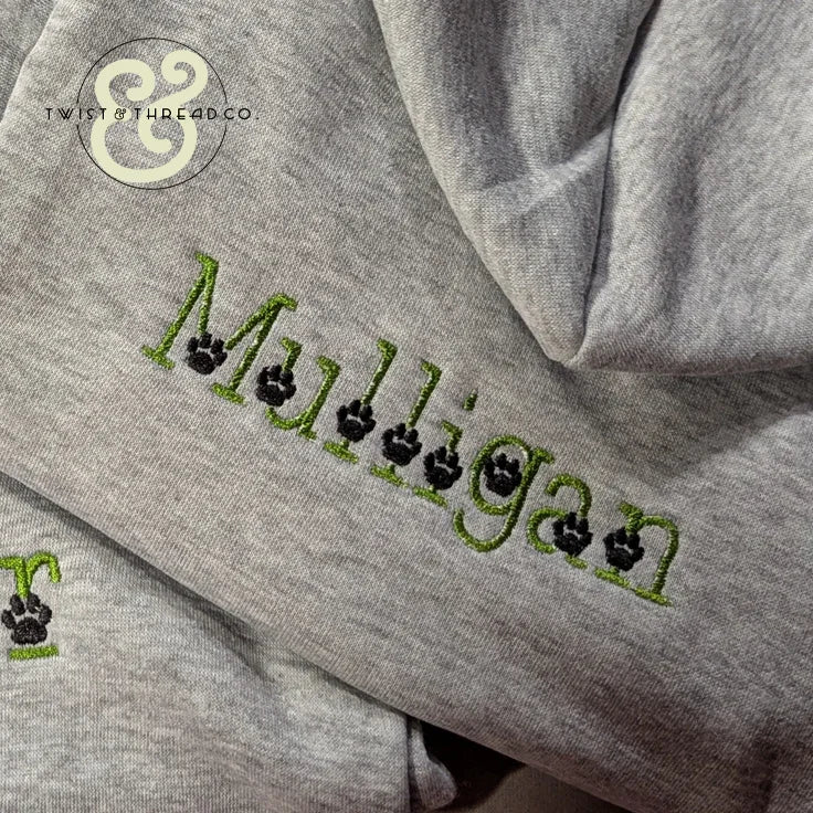 Close-up of gray hoodie with custom green embroidery spelling 'Malligan' featuring black paw prints, from Twist & Thread Co.