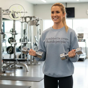 Woman in gym wearing gray sweatshirt with 'dumbbells over dumb people' text holding silver dumbbells