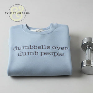 Folded light blue sweatshirt with 'dumbbells over dumb people' text next to a shiny silver dumbbell