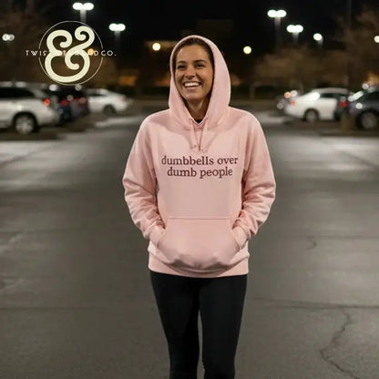 Pink hoodie with text dumbbells over dumb people worn by smiling woman in parking lot at night