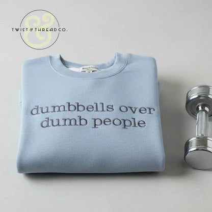 Folded light blue sweatshirt with 'dumbbells over dumb people' text next to a shiny silver dumbbell