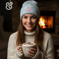 Woman in light blue winter hat and sweater holding mug of hot chocolate by fireplace