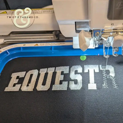 Close-up of embroidery machine stitching white block letters spelling EQUESTR on black fabric for custom equestrian apparel
