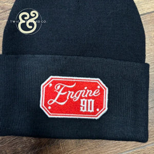 Black knit beanie with red embroidered patch reading 'Engine 90' on wooden surface