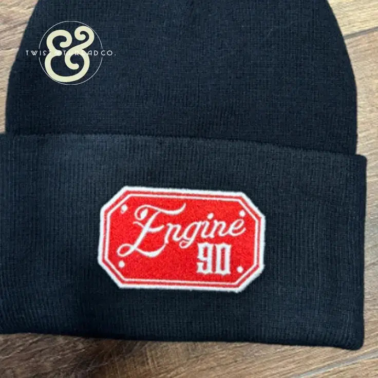 Black knit beanie with red embroidered patch reading 'Engine 90' on wooden surface