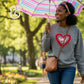 Woman wearing Twist & Thread Co. embroidered heart sweatshirt, holding striped umbrella outdoors