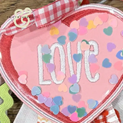 Embroidered pink heart with 'LOVE' text, colorful confetti, and red gingham ribbon from Twist & Thread Co.