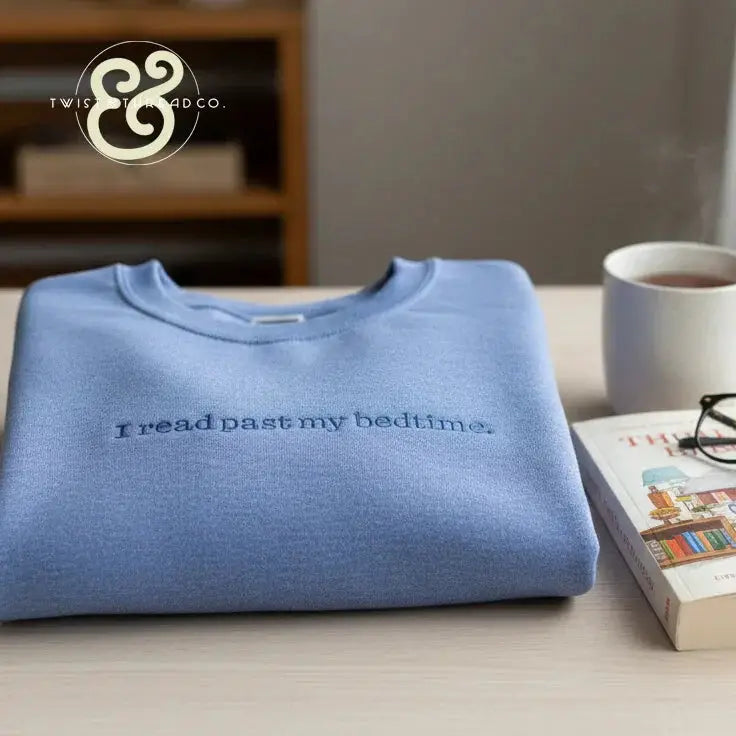 Light blue folded sweatshirt with the phrase 'I read past my bedtime' next to a steaming coffee mug and book