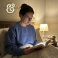 Woman wearing blue sweatshirt reading book in bed by lamp light with stack of books on nightstand