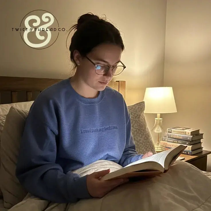 Woman wearing blue sweatshirt reading book in bed by lamp light with stack of books on nightstand