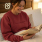 Woman smiling and reading a book in bed wearing a maroon sweatshirt with the text I read past my bedtime