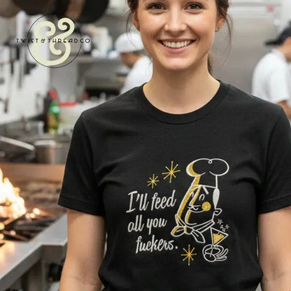 Smiling woman in kitchen wearing black embroidered t-shirt with chef design and text "I'll feed all you" in premium apparel style