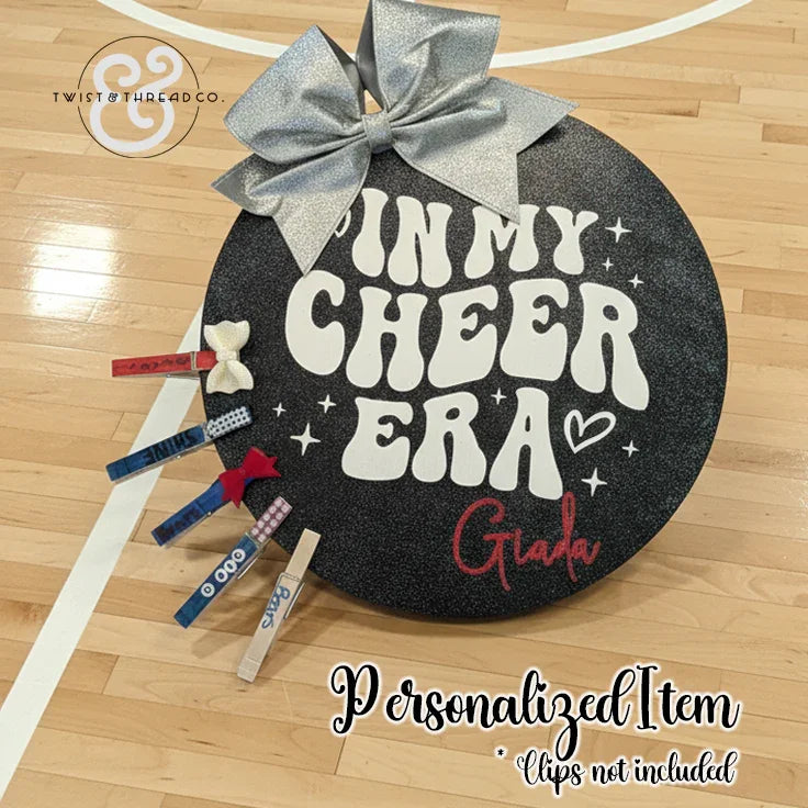 Round black cheer accessory board with silver bow, 'In My Cheer Era' text, on gym floor