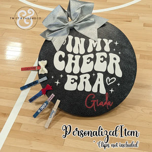 Round black cheer accessory board with silver bow, 'In My Cheer Era' text, on gym floor