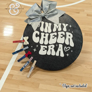 Round black cheer bow holder with silver bow, 'In My Cheer Era' text on gym floor