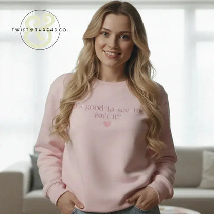 Smiling woman wearing a light pink sweatshirt with text 'It's good to see me, isn't it?' in a cozy living room