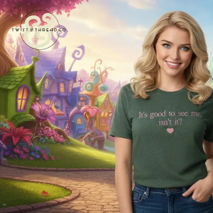 Smiling woman in green T-shirt with text 'It's good to see me, isn't it?' in a whimsical fantasy village backdrop