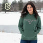 Woman wearing green hoodie with pink text 'It's good to see me, isn't it?' outdoors in snowy winter setting