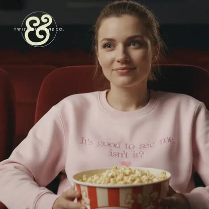 Young woman smiling in theater seat wearing pink sweatshirt with text, holding large popcorn bucket