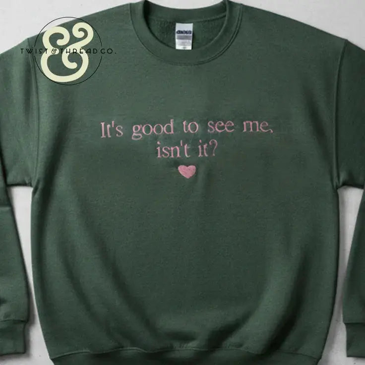 Green sweatshirt with pink text embroidered saying 'It's good to see me, isn't it?' and a heart below