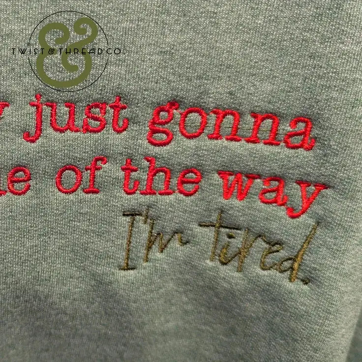 Close-up of gray fabric with embroidered red and brown text reading "I'm just gonna be out of the way I'm tired"