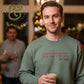 Smiling man wearing green sweatshirt with festive text holding drink at holiday party with blurred lights