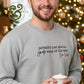 Smiling man wearing gray sweatshirt with 'probably just gonna jingle some of the way I'm tired' text, holding cup, Christmas tree lights
