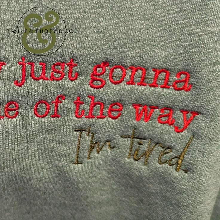 Close-up of gray fabric with embroidered red and brown text reading "I'm just gonna be out of the way I'm tired"