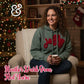 Woman in green Joy hoodie with red yarn, sitting by Christmas tree and fireplace, cozy holiday setting.